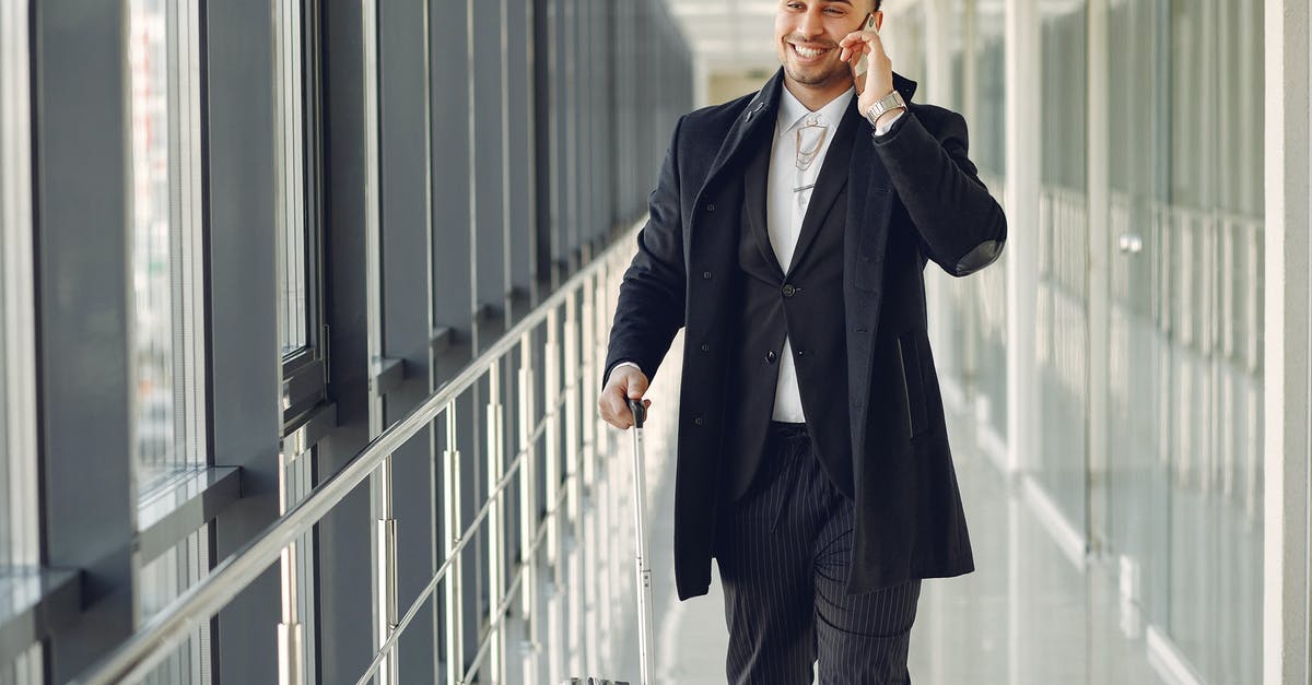 18h layover in Helsinki, can I leave the airport? - Happy ethnic male in formal suit standing in modern hallway while holding suitcase in hand and talking on mobile phone