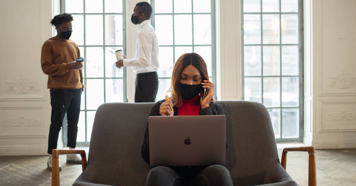 'One laptop per child'- like organisations? - Ethnic female worker in fabric mask with netbook and thumb up speaking on cellphone against male partners with takeaway coffee