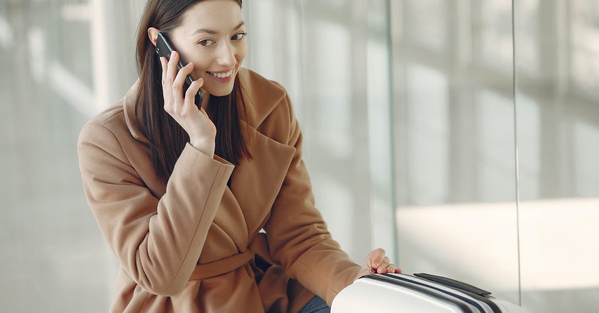 A phone setup for frequent international travel - Young dreamy brunette in beige coat with suitcase having conversation via mobile phone while waiting for flight in airport terminal