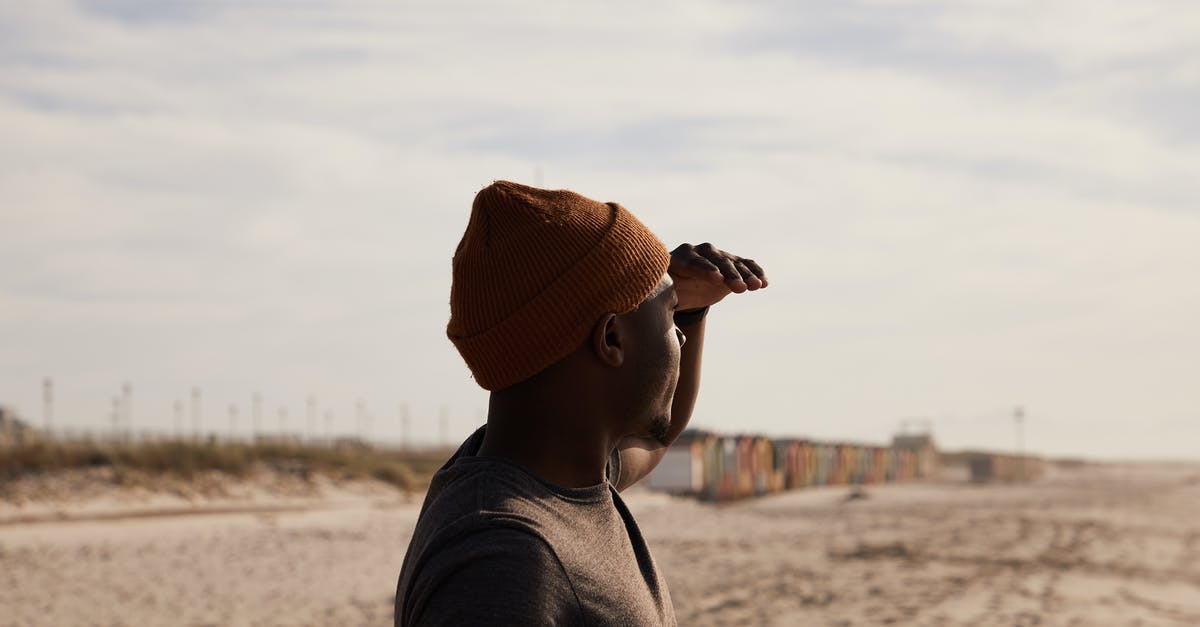 A trip to Patalpani near Indore - Side view of African American male in hat with hand near forehead standing on sandy shore with traces against cloudy sky