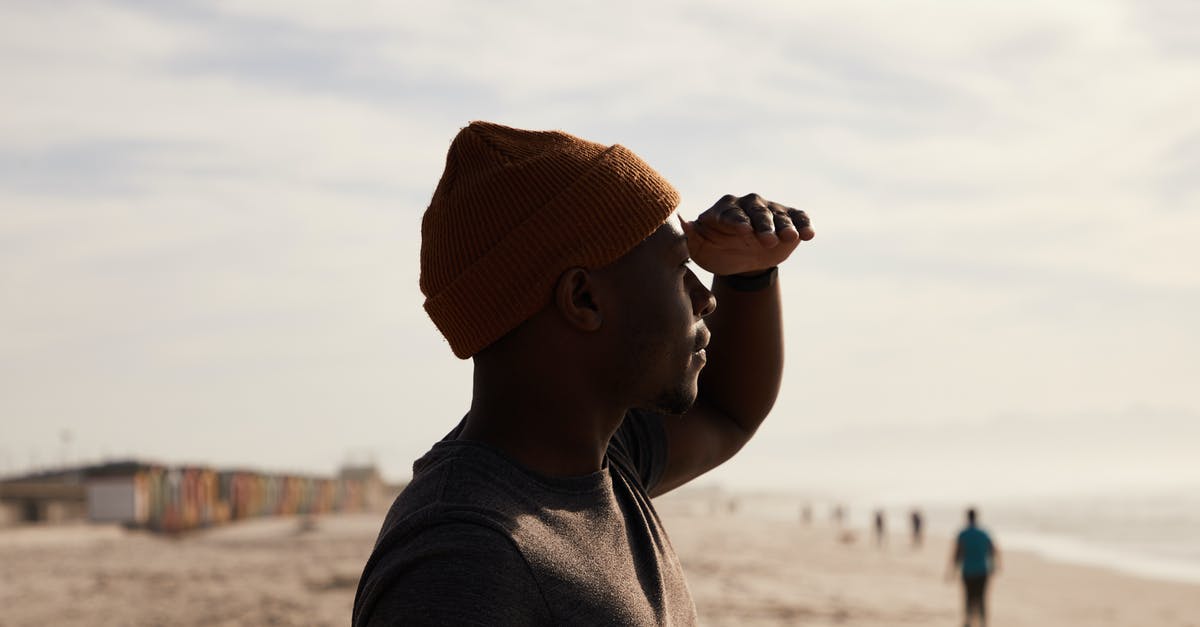 A trip to Patalpani near Indore - Side view of African American male standing with head near forehead on sandy beach near sea with blurred people against cloudy sky