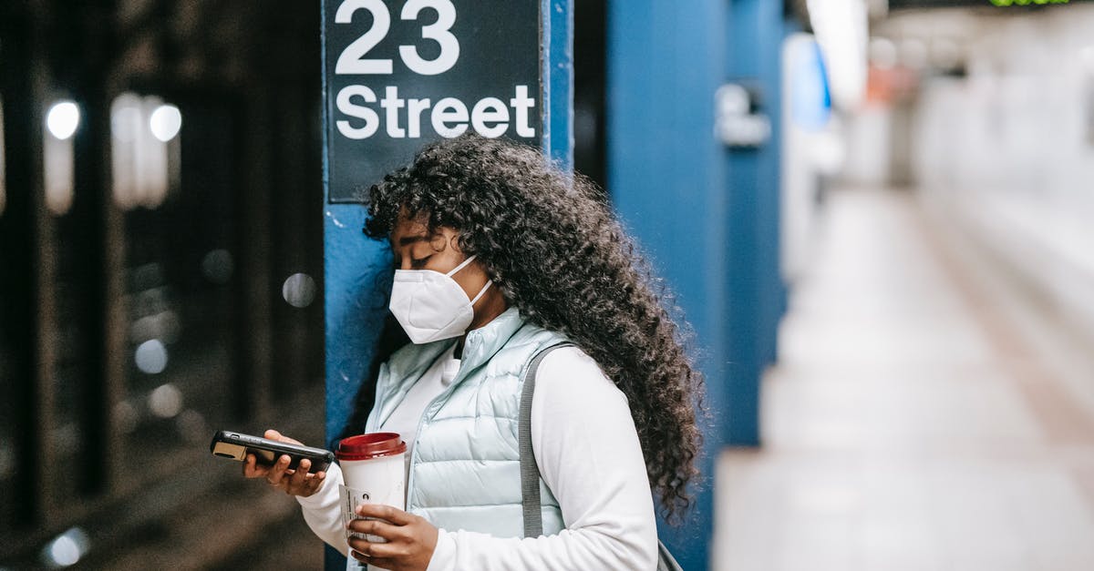 About visa transit lisbon Portugal/ usa - Focused African American female wearing casual outfit and protective mask standing on New York underground platform with takeaway coffee and using mobile phone