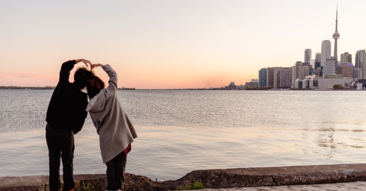 Accompanying friend for a Canada visa? - Back view full body couple in casual wear forming heart with hands while standing on seaside and admiring scenery of Toronto cityscape