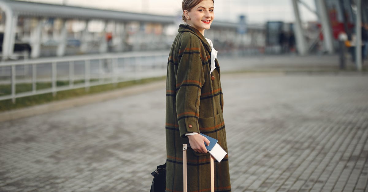 Adding a connecting flight to an existing ticket - Side view of positive young woman in warm clothes smiling at camera while carrying luggage with passport walking along airport terminal