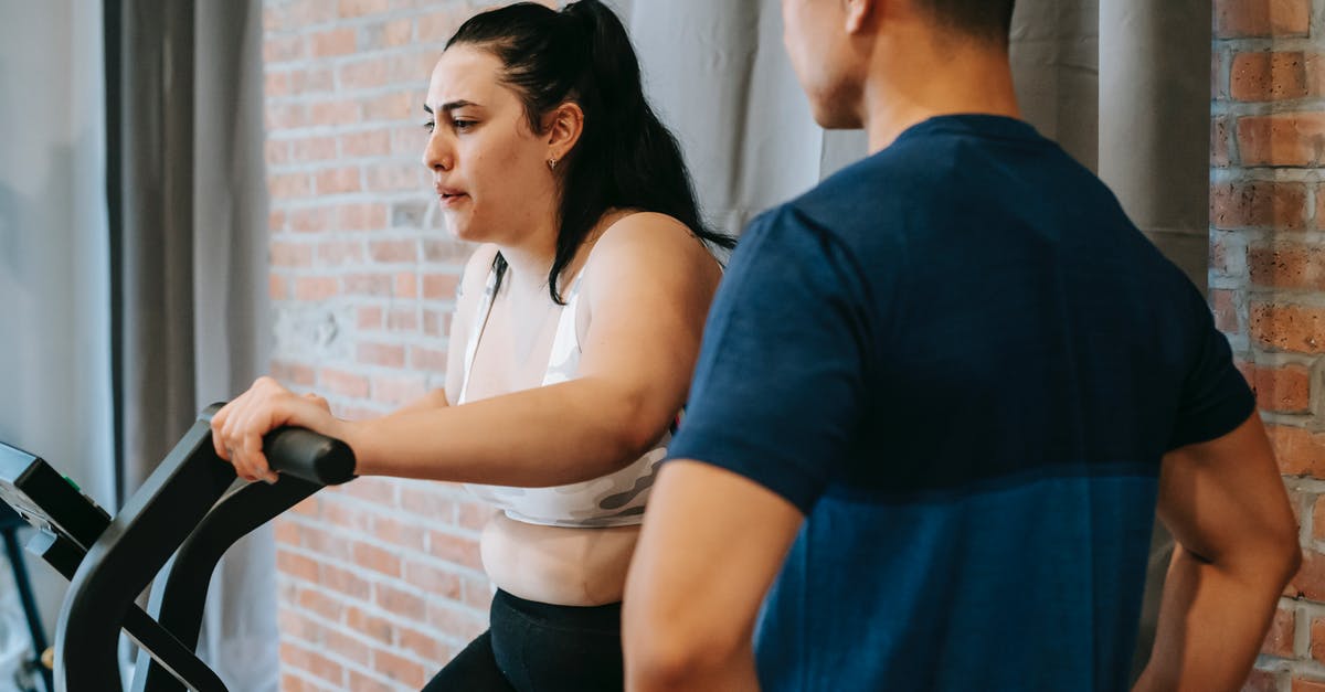 Additional weight when taking a bike with Air Brunei - Side view of serious concentrated young obese female doing weight lose exercise on cycling equipment during cardio training with crop ethnic male personal coach