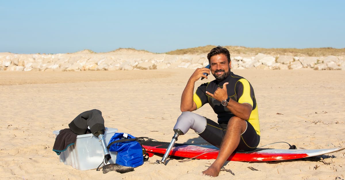 Airlines with the most Surfboard friendly baggage policy & fees pricing? - Male with prosthesis looking at camera sitting on surfboard while having conversation on smartphone and showing shaka