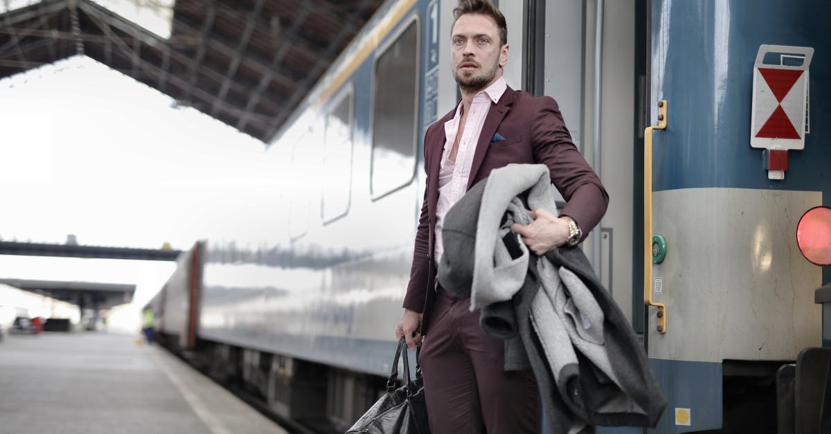 Airport transit in Amsterdam - Serious stylish bearded businessman in trendy suit holding bag and coat in hands standing near train on platform in railway station and looking away