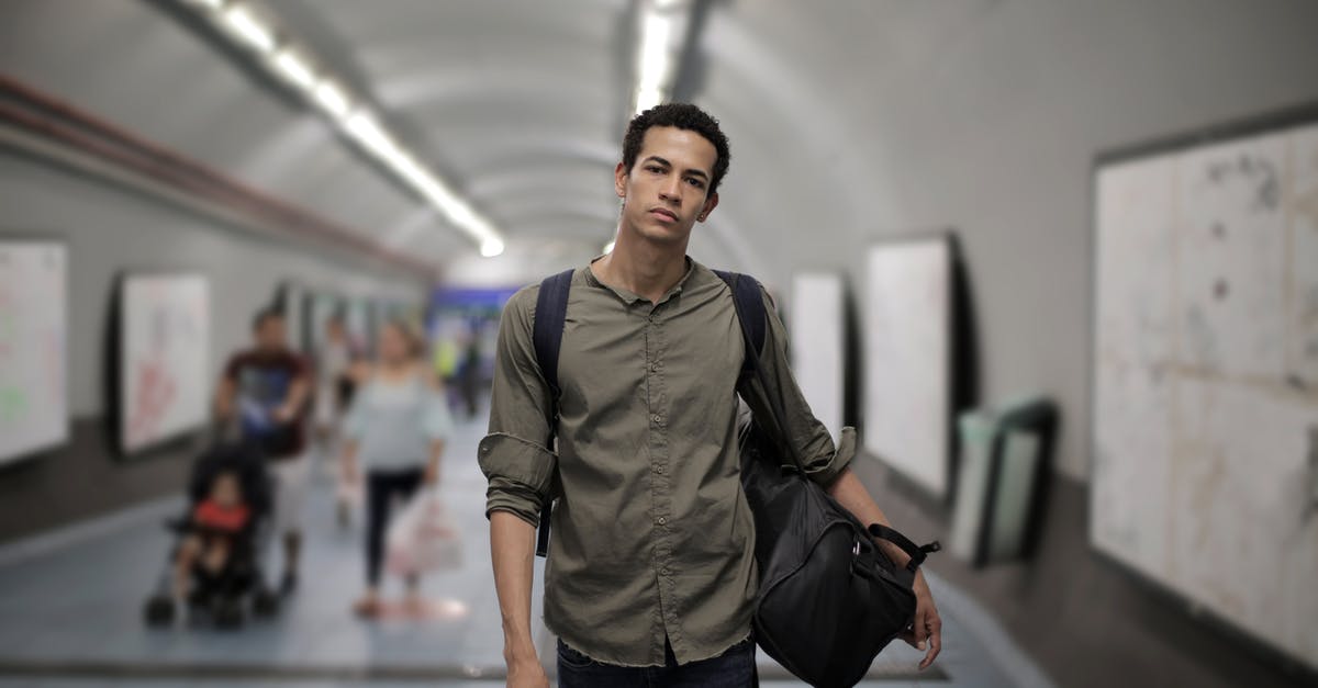 Airport transit US to Asia [duplicate] - Calm young African American male in casual clothes with big black bag and backpack looking at camera while walking along corridor of underground station against blurred passengers