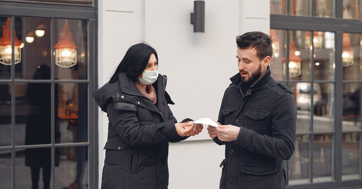 Any suggestions to ensure safety while hitchhiking? - Careful female volunteer in black down jacket suggesting protective facial mask for bearded male pedestrian on city street in cold season during coronavirus pandemic