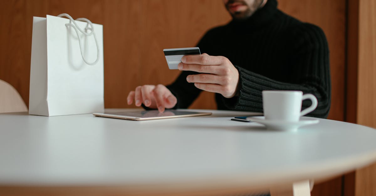 Applying for a Schengen visa using spouse's bank statement - Crop focused bearded young man using tablet while doing online purchase with credit card sitting at table in modern cafe with coffee cup and shopping bag