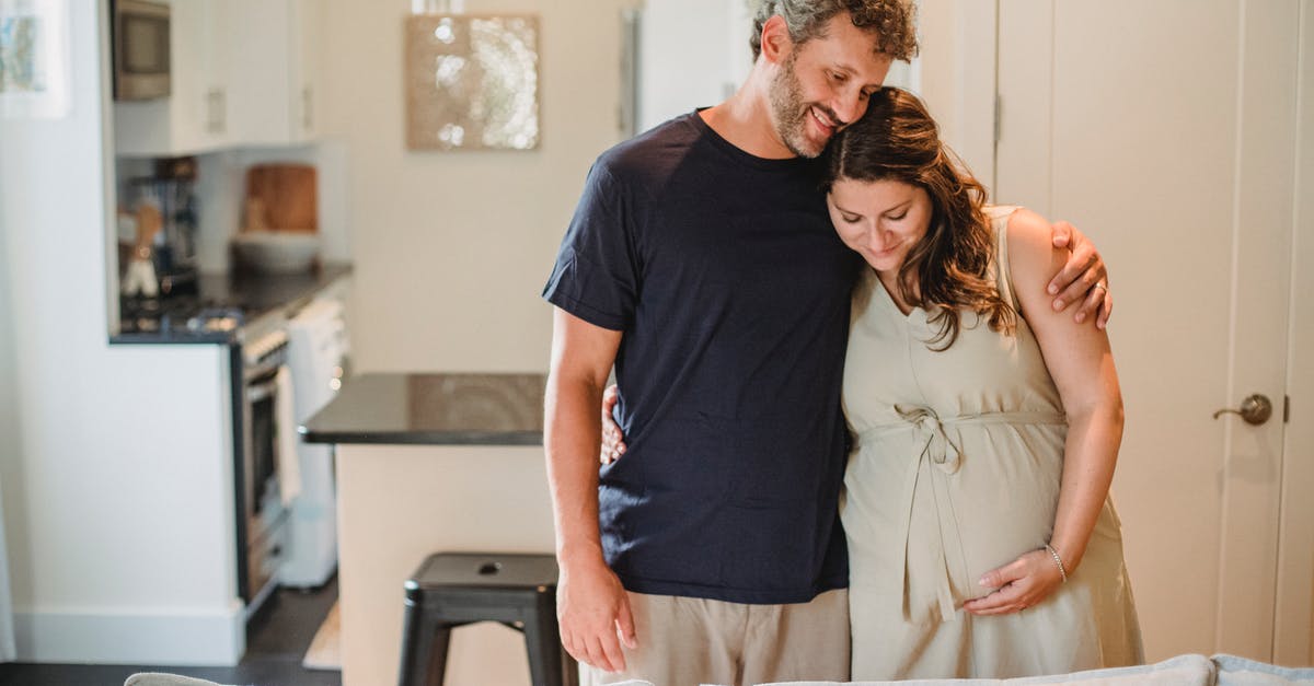 Applying for belgium visa with pregnant wife [closed] - Smiling bearded man with curly gray hair embracing happy pregnant wife in beige dress in room of modern apartment