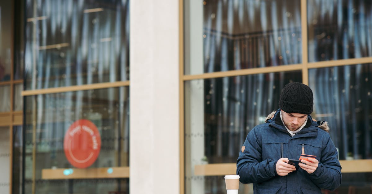 App/service that suggests which payment card is most convenient? - Focused young male in warm outerwear and hat standing near modern building with takeaway coffee and using smartphone while making online shopping with credit card