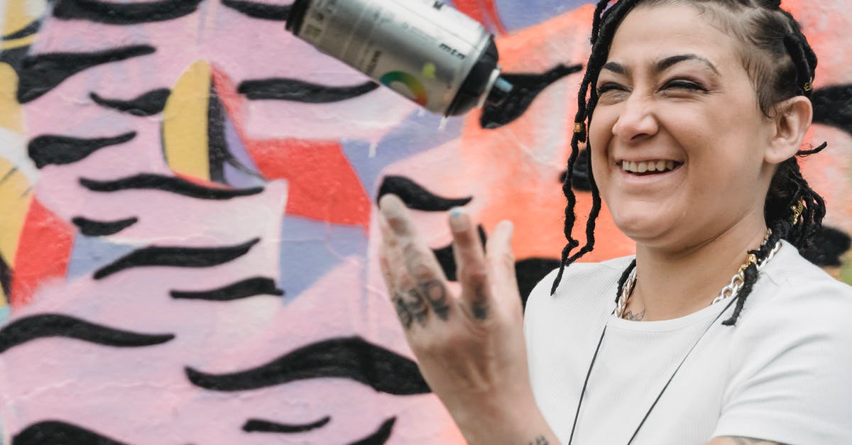 Are aerosol cans allowed and safe, in checked luggage? - Happy ethnic female artist with braided hair tossing up spray paint can while standing near wall with creative graffiti on street