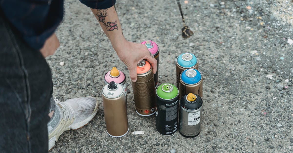 Are aerosol cans allowed and safe, in checked luggage? - Crop faceless tattooed artist taking paint bottle from heap of multicolored spray cans placed on ground on street of city