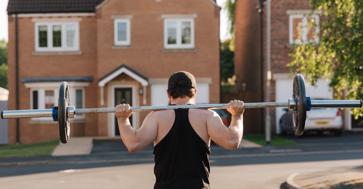 Are there any Squat houses in Paris? - Back view of anonymous fit bodybuilder in cap squatting with barbell while working out on pavement against houses in sunlight