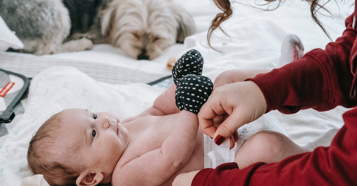 Are there baby changing rooms at Delhi airport? - Crop anonymous mother putting on diaper on adorable infant baby wearing anti scratching mittens while lying on comfortable couch near hairy dog in light room