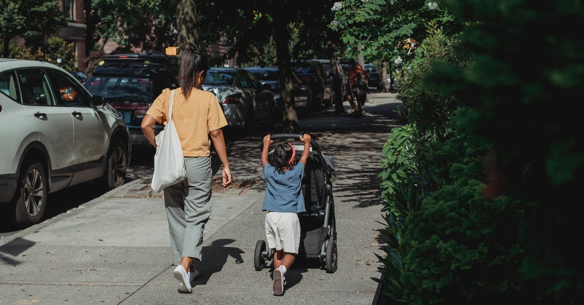 Arranging temporary childcare during a conference in an unfamiliar city - Full body back view of anonymous ethnic woman with bag walking on sidewalk near girl pushing baby stroller on sunny street