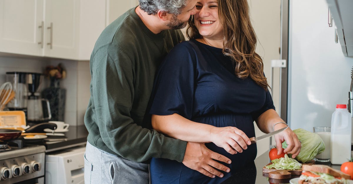 B2 tourist visas for my future wife and her parents - Cheerful pregnant wife in blue dress cooking sandwiches for breakfast while smiling and looking at husband embracing belly in modern kitchen