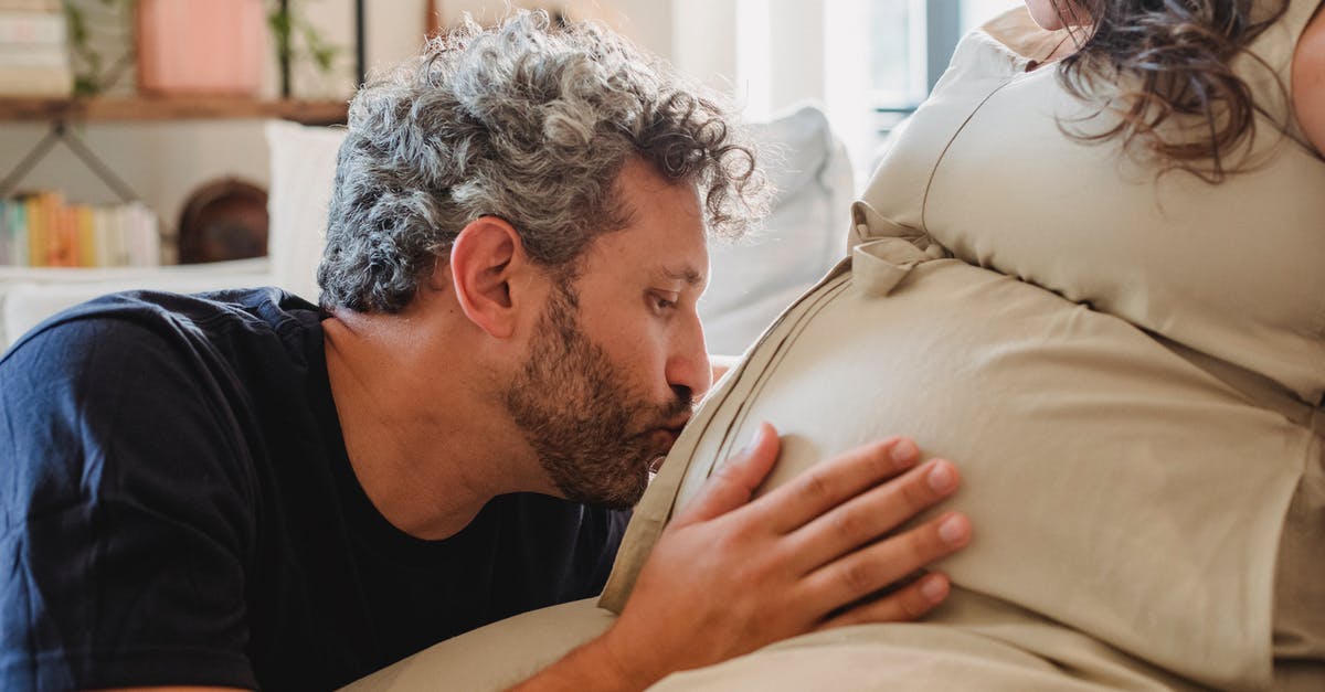 B2 tourist visas for my future wife and her parents - Side view of loving adult husband gently touching and kissing pregnant tummy of crop wife sitting on couch in light apartment