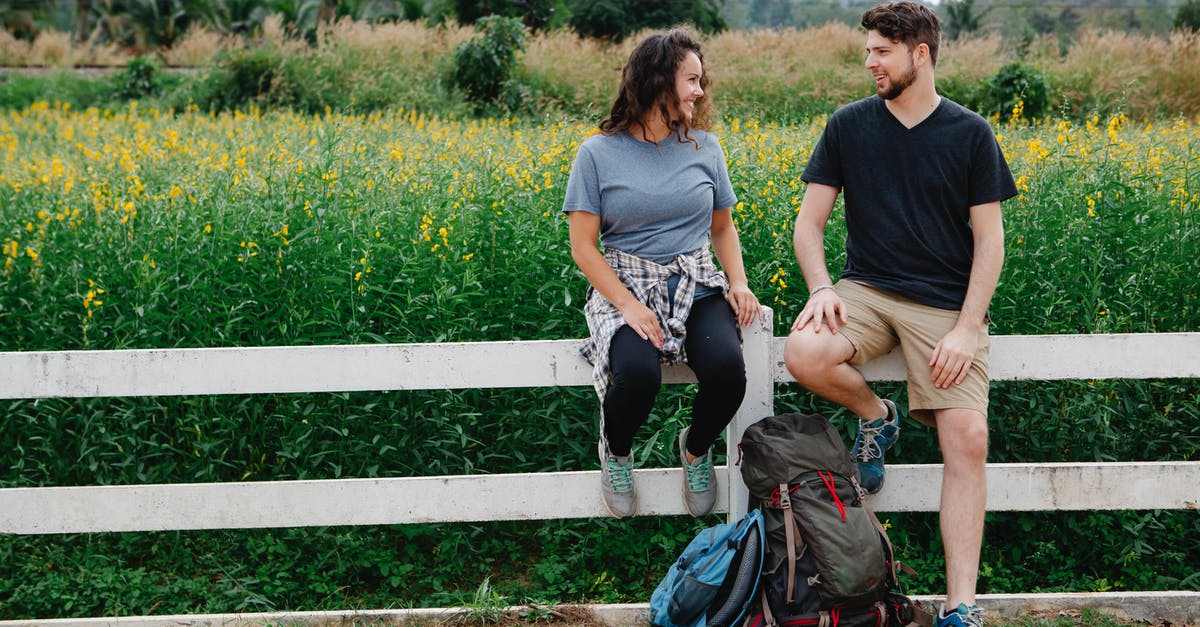 Backpack trip to Europe [closed] - Romantic young couple resting on wooden fence near blooming lush meadow and chatting during hiking trip in countryside