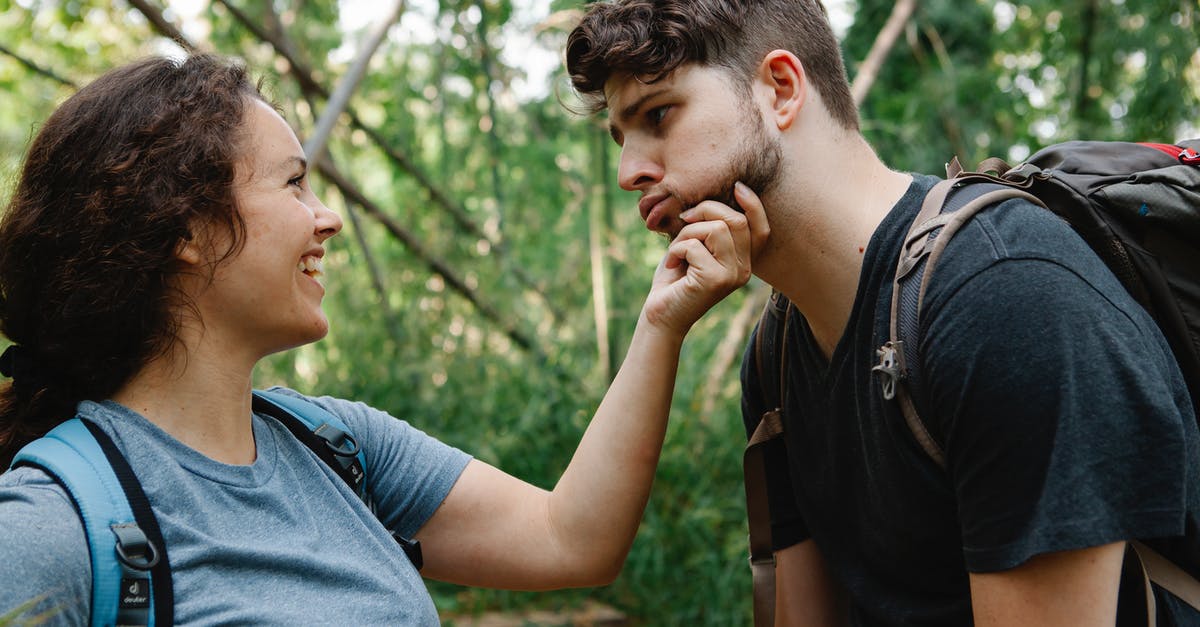 Backpack trip to Europe [closed] - Side view of happy young woman smiling and touching face of tired boyfriend making grimace while standing together in green forest during hiking trip