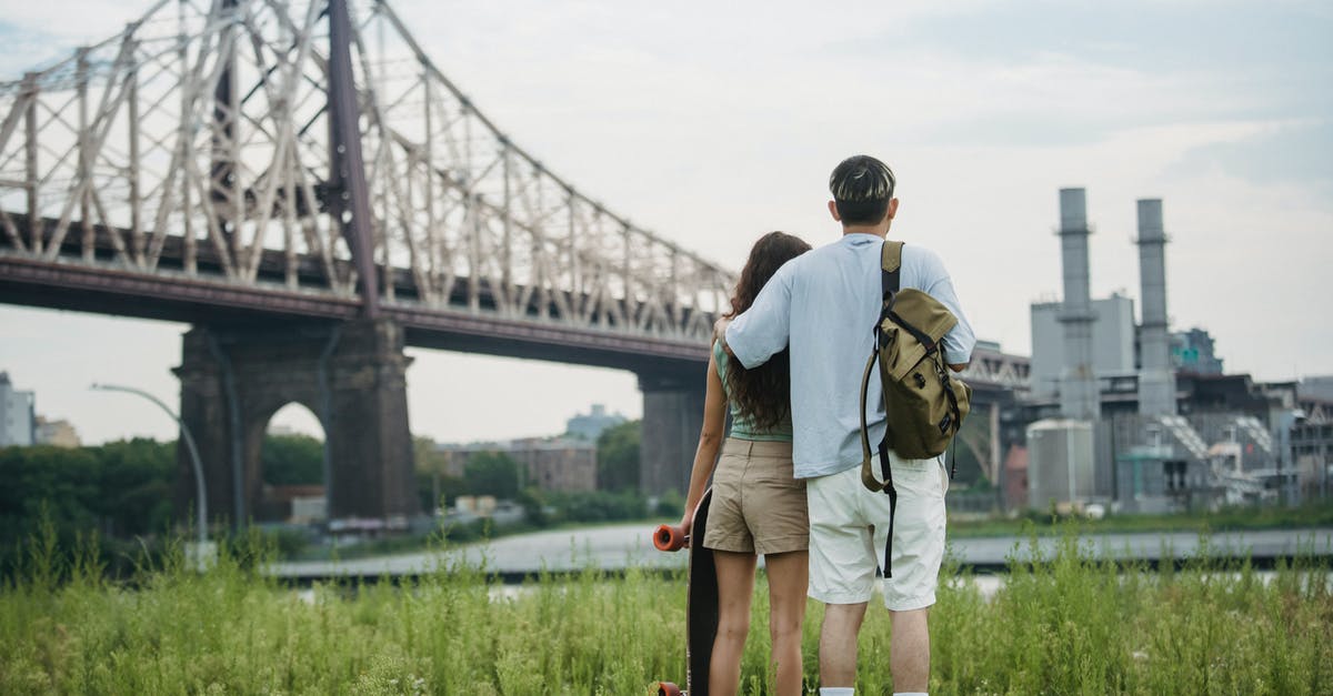 Backpacking with visa B2 in USA - Back view of unrecognizable young male tourist in casual clothes with backpack cuddling girlfriend with skateboard in hand while standing on grassy ground near Brooklyn Bridge and admiring city