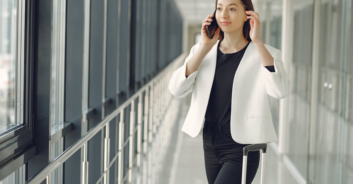 (Baggage) interlining/transit formalities between BA and AI at LHR - Positive young businesswoman in stylish formal wear with suitcase talking on smartphone and standing in modern airport corridor while waiting for flight