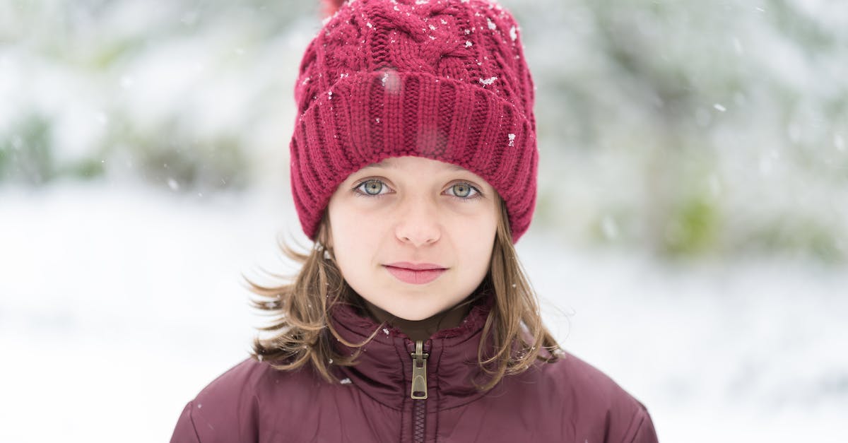 Bath to London on Christmas Day - Calm young girl with short brown hair and light eyes in winter down jacket and hat with pompom standing on street with falling snowflakes in winter park