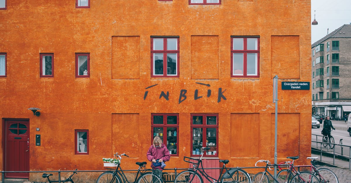 Bicycle parking at Narita airport - Woman parking bicycle near metal railing against colorful orange stone house with shop downstairs located in Copenhagen Denmark on cloudy day