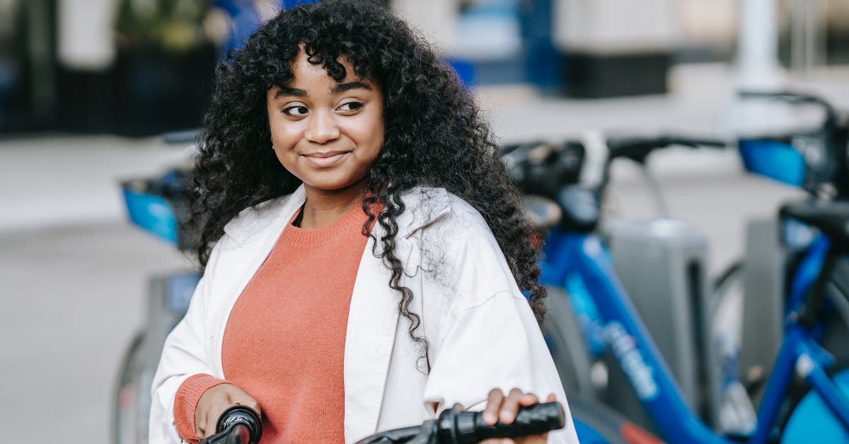 Bike sharing apps-rent cycle in one and back in other city - Smiling African American female cyclist wearing casual outfit standing with bicycle near bicycle sharing station on modern city street