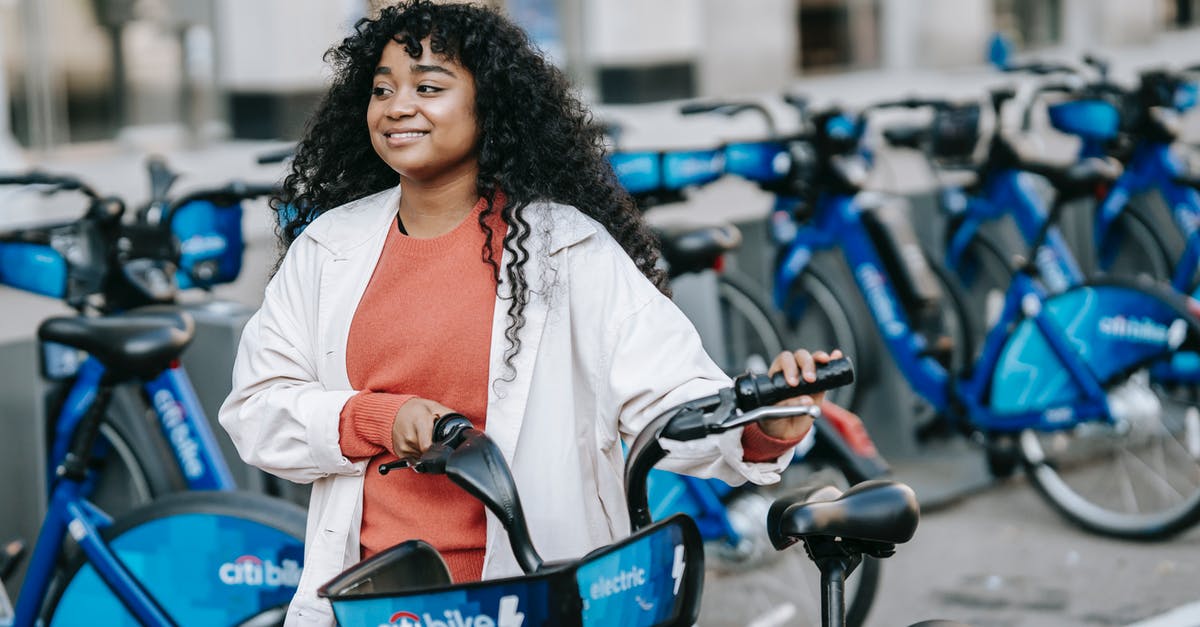 Bike sharing apps-rent cycle in one and back in other city - Cheerful African American female bicyclist in casual clothes renting bicycle in bicycle sharing station on city street and looking away contentedly