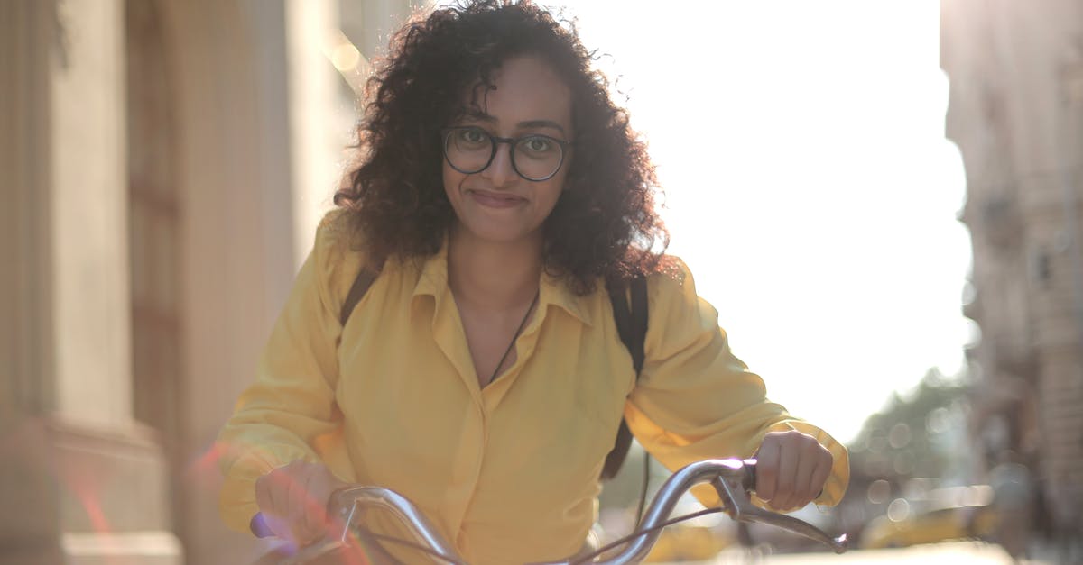 Bike transport from Haridwar to Bangalore - From below cheerful young curly woman in glasses with backpack smiling at camera while riding bicycle along street in sunny day