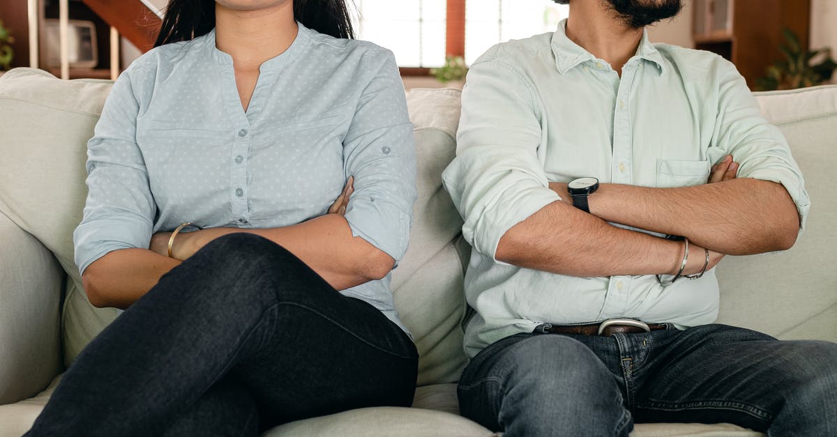 Booking flight tickets together or in separate parts? - Crop faceless young ethnic couple in casual wear sitting on couch with arms crossed keeping silence after scandal at home