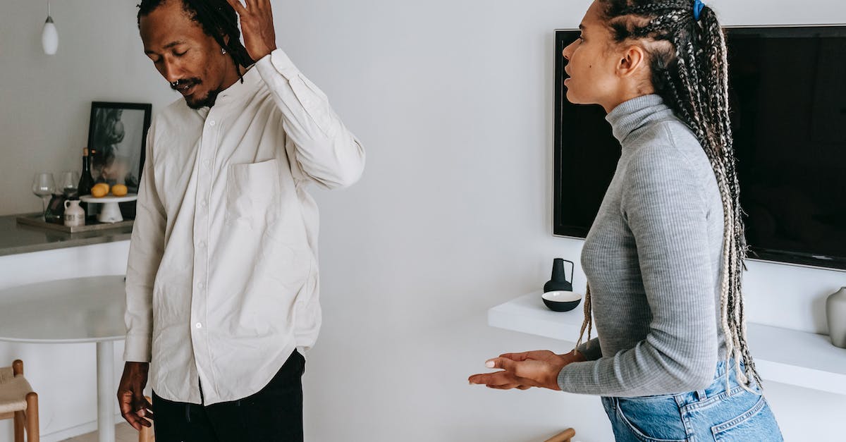 Booking flight tickets together or in separate parts? - Tired young ethnic man with dreadlocks in casual outfit gesticulating while having argument with wife at home