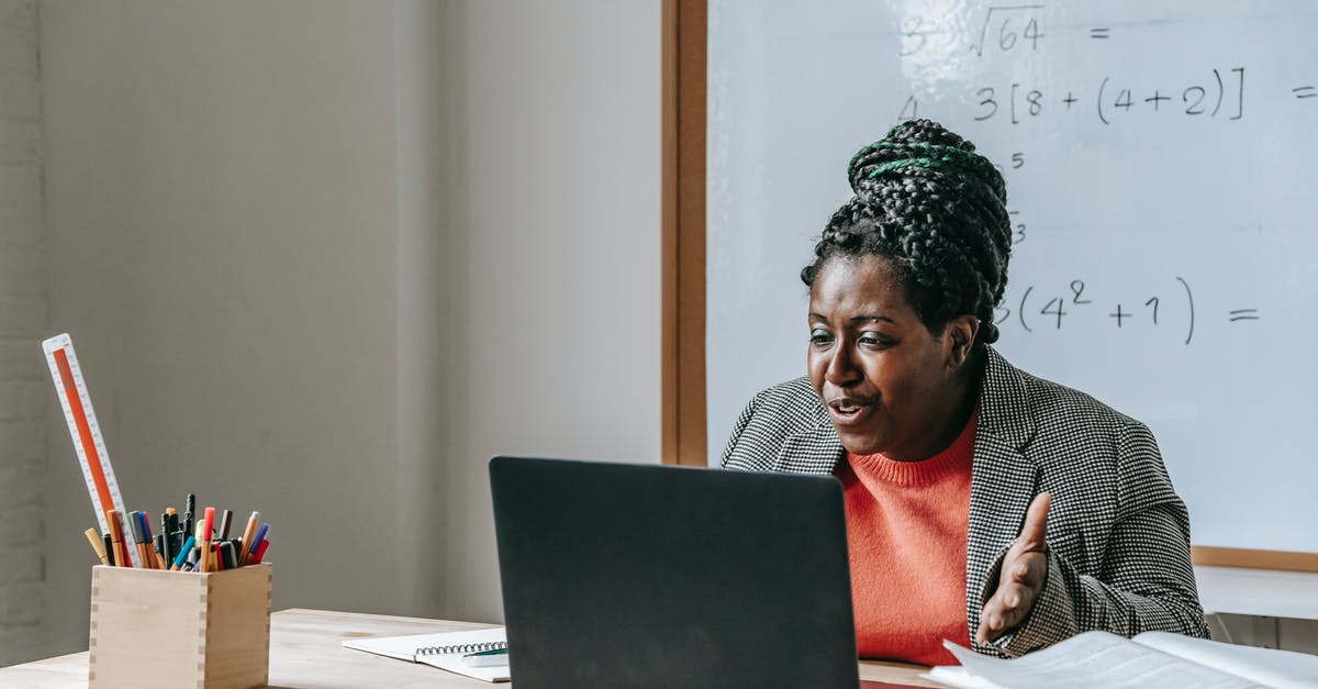 Booking Reference Number - African American female teacher talking during class on netbook while sitting at desk with books and stationery in classroom