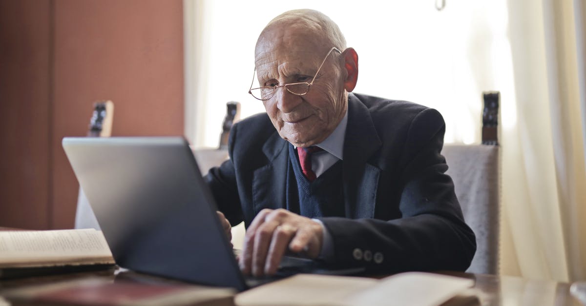 Booking.com: can multiple people manage one booking? - Focused elderly man in formal black suit and eyeglasses using laptop while sitting at wooden table with books in light room