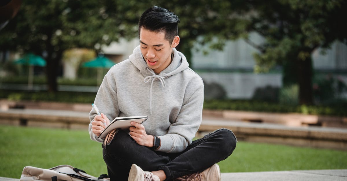 Border crossing on Curonian Spit - Full body of concentrated Asian male in gray hoodie and ripped black jeans sitting on stone border with legs crossed and writing in notepad