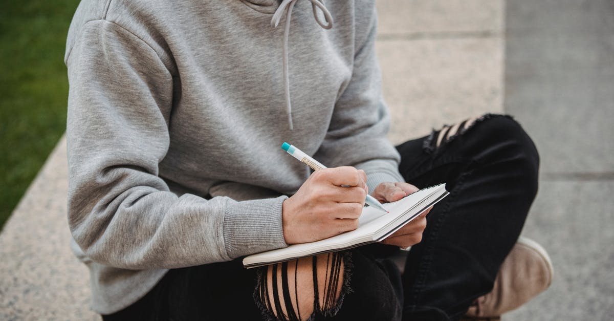 Border crossing on Curonian Spit - Crop unrecognizable male in gray hoodie and ripped black jeans sitting on stone border with legs crossed and taking notes in notebook