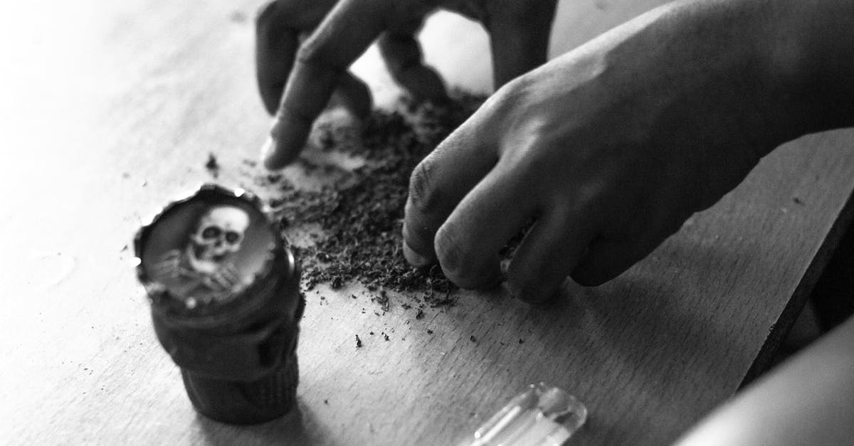 Bringing a medicine containing Cannabis Sativa from India to the Netherlands - From above of black and white crop anonymous male rolling up joint with weed at table with cup with skull