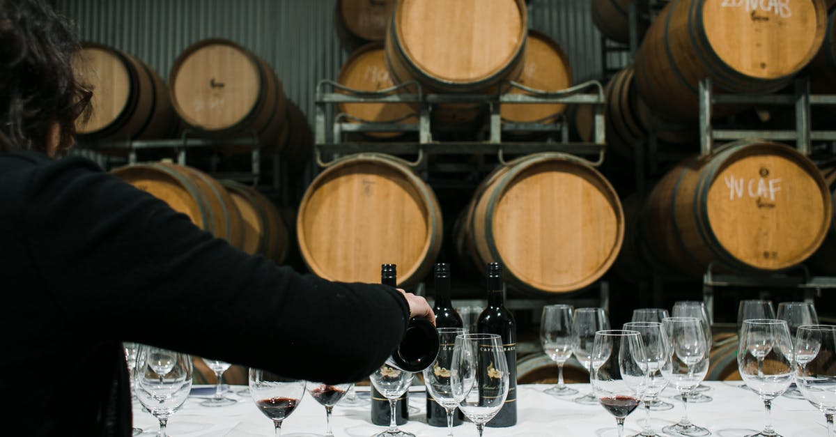 Bringing extra alcohol back to the US: what will it cost? - Back view of anonymous sommelier pouring wine from bottle into wineglass while standing at table with glassware in winery against barrels