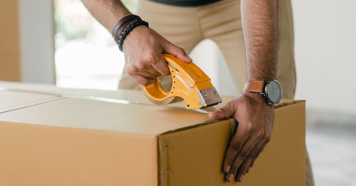 Bringing work goods across border - Crop faceless young male with wristwatch using adhesive tape while preparing cardboard box for transportation