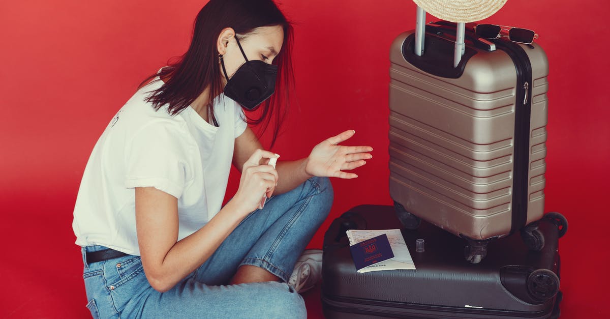 Bug spray on my suitcase - Side view of young woman in casual clothes and medical mask sitting near luggage with passport and tickets while disinfecting hands on red background