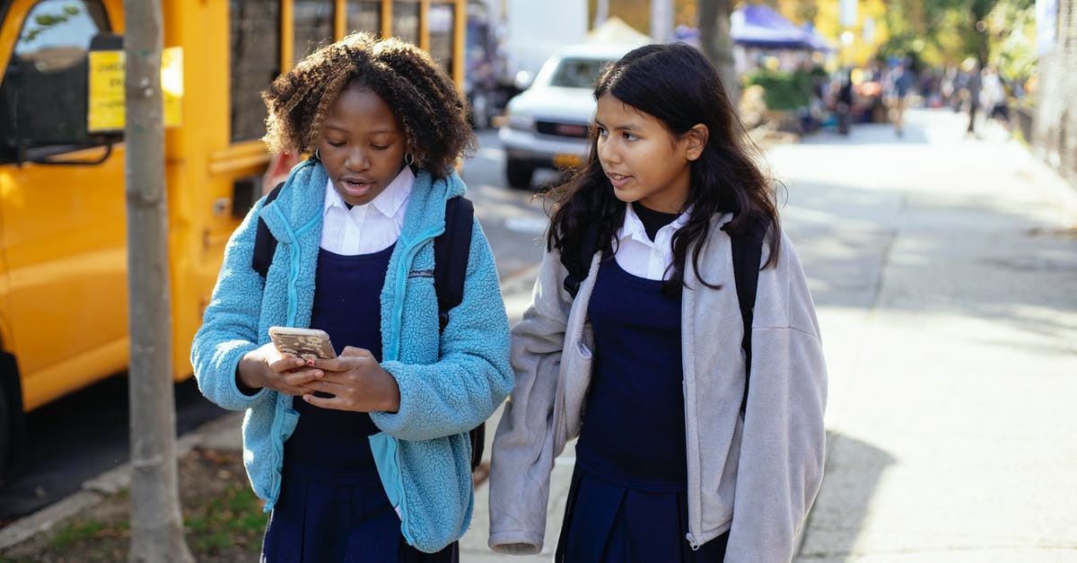 Bus connection from Jesenik to Vidnava, Czech Republic - Cheerful multiracial girls classmates with backpacks browsing smartphone while walking together on sunny autumn street near yellow school bus