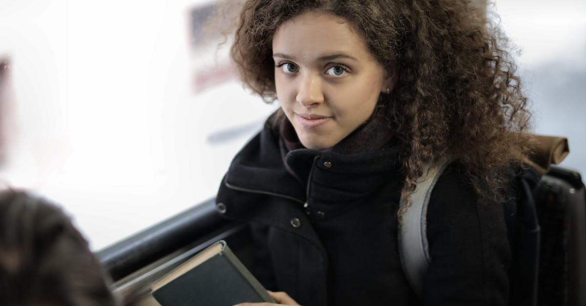 Bus travel from Salgótarján to Nagymaros on Easter Sunday - From above view of positive young lady with curly hair in casual warm coat sitting with book on passenger seat near window in bus and looking at camera