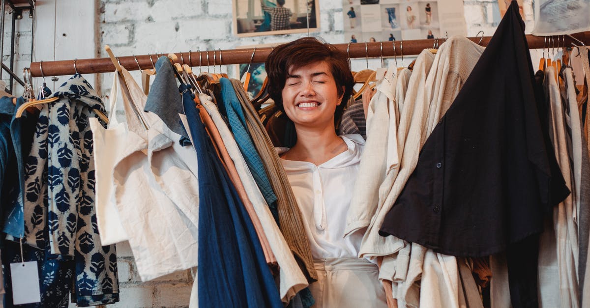 Buy airfares now or after the sale? [closed] - Cheerful Asian female customer standing among hanging clothes in store and smiling