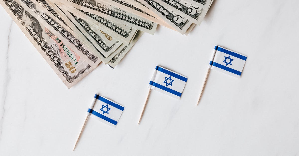 Buying a flight ticket from a different country - Top view of bundle of different nominal pars dollars and Israeli flags on toothpicks placed on white surface of marble table