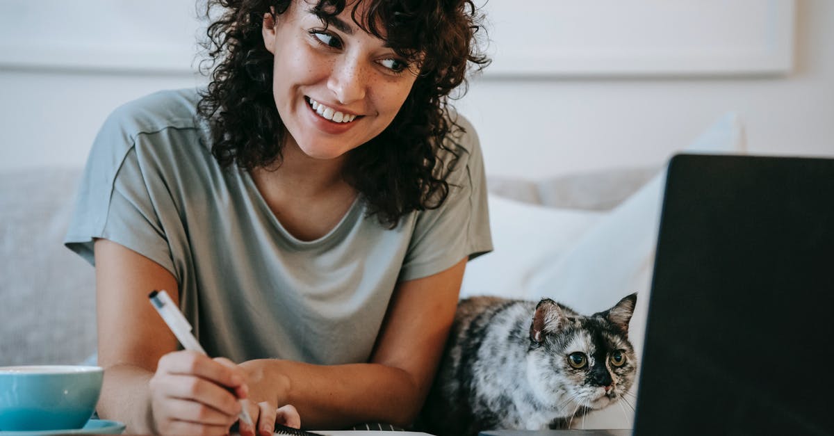 Buying domestic flights in India online - Crop young positive female smiling and taking notes in organizer while cat watching on netbook at table at home