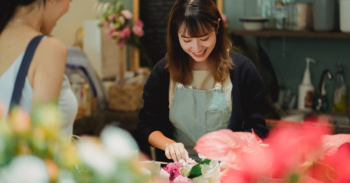 Buying Interrail pass abroad without passport - Content Asian female florist wearing light green apron composing tender bouquet for positive female customer while working at counter in floristry shop