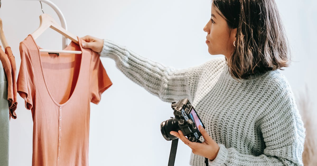 Camera store near Dubrovnik, Croatia? - Side view of young female in sweater selecting shirt from hanger for photoshoot while holding camera in light studio