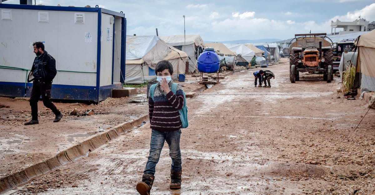 Camping safety in the Everglades in May - Full body of ethnic boy in protective mask strolling on dirty path along shabby tents in poor district of countryside during pandemic